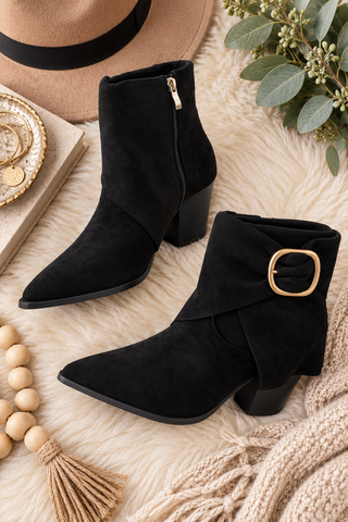 A pair of black suede ankle boots with pointed toes, block heels, and gold-tone buckle details, styled on a cream faux fur rug with a hat, beads, and greenery.