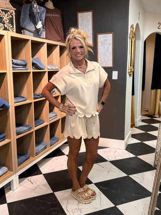 A woman modeling a cream-colored short-sleeve polo shirt and matching shorts with scalloped hems, standing in a boutique with a black and white checkered floor and wooden display shelves.