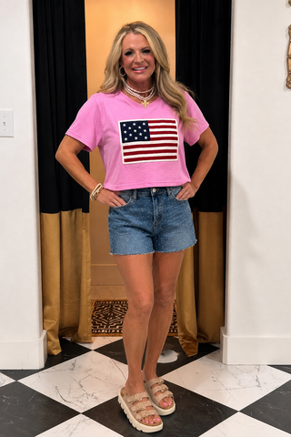 Woman wearing a pink t-shirt with an American flag design and denim shorts indoors.