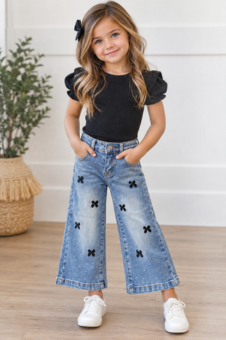 A young girl stands indoors wearing a black short-sleeve top with ruffled shoulders and light blue wide-leg denim jeans decorated with black bow details. She is wearing white sneakers and has a black bow in her hair, standing on a light wood floor in front of a white paneled wall.