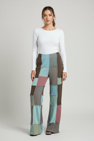 A model wearing a white long-sleeve top and high-waisted patchwork pleated trousers in shades of brown, teal, pink, and gray, standing against a light gray studio background.