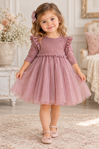A young girl with blonde wavy hair wearing a dusty rose pink tulle dress with ruffled sleeves and a bow headband, standing in a beautifully decorated room.