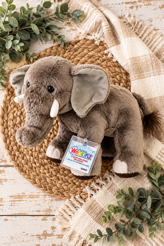 A gray plush elephant toy with white tusks sits on a round woven placemat, surrounded by a beige plaid throw blanket and green eucalyptus leaves. A hangtag reading 'Weaning' is attached to the toy.