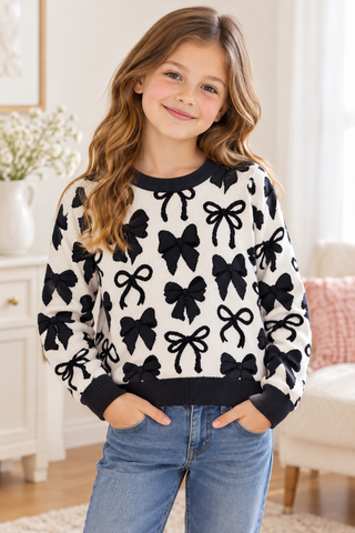 A young girl with wavy light brown hair smiling while wearing a black and white knit sweater with a bow pattern, paired with blue jeans, standing in a bright, modern living room.