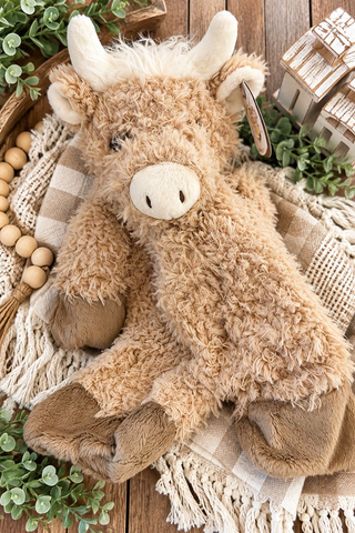 A plush stuffed animal shaped like a fluffy brown cow or ox, posed on a woven blanket with decorative greenery and wooden beads around it on a rustic wood surface.