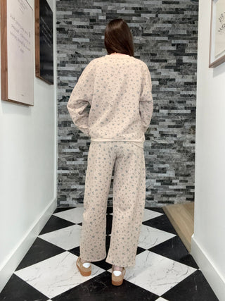 A woman viewed from behind wearing a matching beige pajama set with a small blue floral pattern, standing in a hallway with a black and white checkered floor and a gray stone accent wall.
