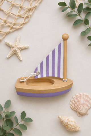 A wooden toy sailboat with a purple and white striped sail and a purple hull, styled on a light beige background with seashells, a starfish, green leaves, and a net.