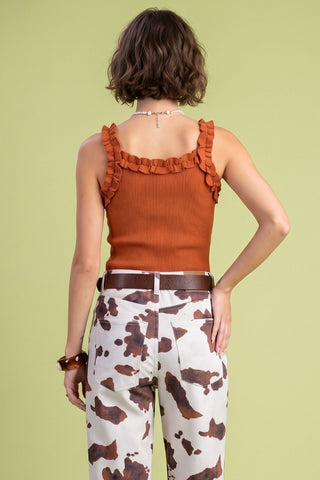 A woman shown from the back wearing a rust-orange ribbed tank top with ruffled straps and white cow-print pants, standing against a solid light green background.