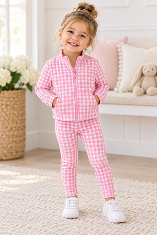 A young girl stands in a bright room wearing a pink and white gingham zip-up jacket and matching gingham pants, paired with white sneakers.
