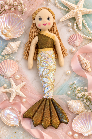 A mermaid doll with long blonde braided hair, a brown top, and a glittery gold and iridescent tail, styled on a pastel marble background with seashells, starfish, pearls, and pink ribbon.
