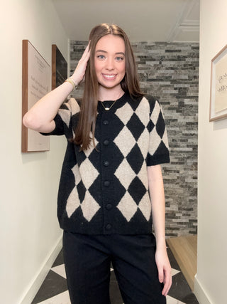 A woman modeling a short-sleeve black and beige argyle knit cardigan with dark buttons, paired with black pants, standing in an indoor hallway with a gray stone accent wall behind her.