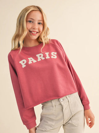 A young blonde woman wearing a dusty rose cropped sweatshirt with 'PARIS' printed in white across the chest, paired with light beige jeans, against a solid cream background.