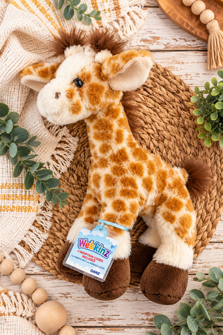 A plush giraffe toy with a patterned brown and cream body, lying on a woven mat with a tag attached. The tag reads 'Webkinz' and 'GANC'.