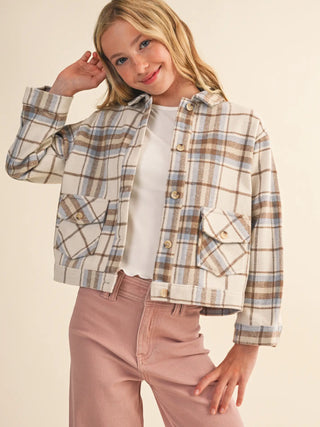 A young girl wearing a cropped, long-sleeve plaid button-up jacket in beige, brown, and light blue over a white t-shirt and dusty pink pants, posed against a solid cream background.