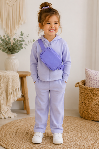 A young girl stands in a cozy room wearing a light lavender two-piece tracksuit with a matching purple crossbody bag, white sneakers, and a purple hair tie.