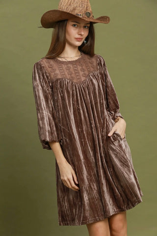 A woman wearing a brown velvet dress with a sheer embroidered yoke and a matching brown cowboy hat, standing against a solid olive green background.
