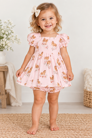 A toddler girl stands barefoot on a woven rug wearing a white smocked dress with a brown and tan dog print, matching shorts, and a cream bow in her hair. The background is a bright, neutral room with a wooden side table and flowers.