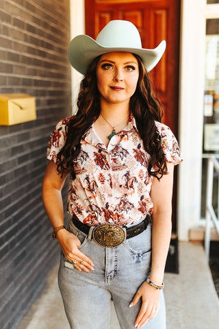 Woman wearing a cowboy hat, patterned shirt, and jeans standing against a brick wall.
