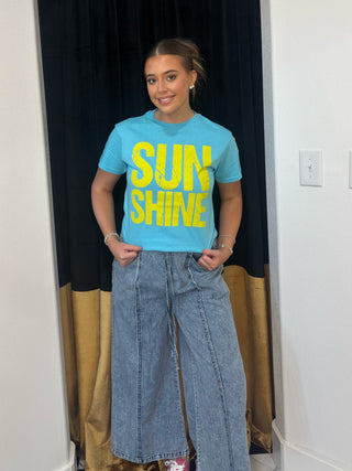 Person wearing a blue t-shirt with 'SUN SHINE' text in yellow, standing in a room with black and gold curtains.
