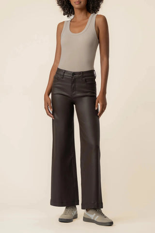 A model wearing dark brown wide-leg faux leather pants, a light beige tank top, and gray sneakers, standing against a solid cream background.