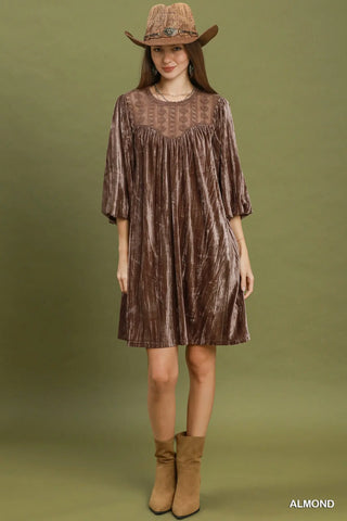 A woman modeling a brown textured midi dress with three-quarter sleeves and a sheer embroidered yoke, styled with a brown hat and tan boots against an olive green studio background.