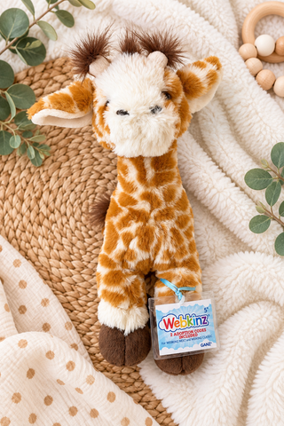 A plush giraffe toy with brown and cream coloring, styled on a woven mat and cream blanket with baby accessories. A hangtag for 'Webkinz Giraffe Shoes' is attached to its leg.