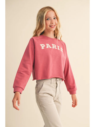 A young girl with blonde hair smiling, wearing a dusty rose pink cropped sweatshirt with 'PARIS' printed in white across the chest, paired with light beige pants, against a solid beige background.