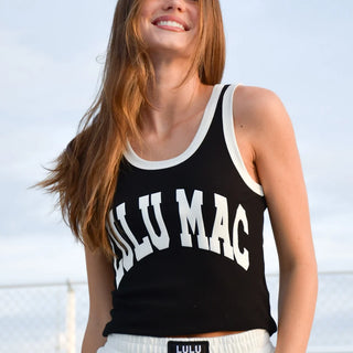 A woman wearing a black cropped tank top with white trim and 'LULU MAC' printed in white arched letters across the front, paired with white sweatpants. The background shows a soft-focus outdoor scene with a chain-link fence and sky.