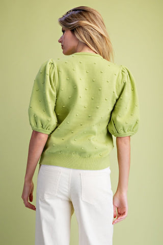 A woman shown from the back wearing a light lime green knit sweater with textured 3D puff sleeves and white pants, set against a solid light green background.