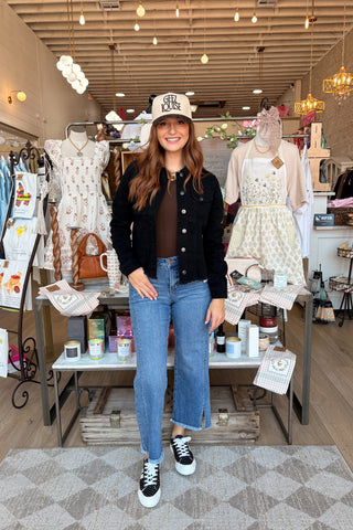 A woman stands in a boutique wearing a black denim jacket, brown top, blue jeans, and a white baseball cap with 'ELK' embroidered on it. The shop interior has mannequins with dresses, hanging lights, and display tables.