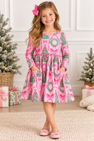 A young girl with a pink bow in her hair stands in a holiday-themed room wearing a pink long-sleeve dress with a green and pink Christmas print, including bows, wreaths, and pine trees. She is standing on a beige rug with Christmas trees and wrapped gifts in the background.