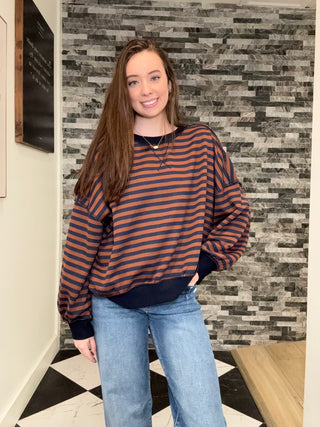A woman wearing a navy and rust orange striped crewneck sweatshirt with balloon sleeves, paired with light blue jeans, standing in front of a gray stone-tile wall.