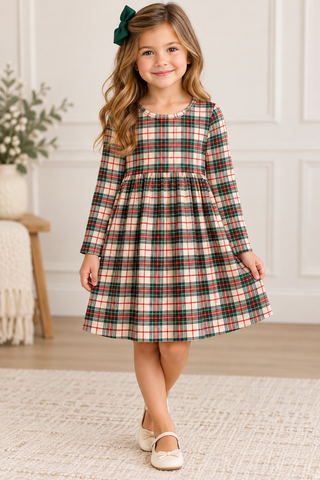 A young girl with long wavy brown hair wearing a green bow headband, a long-sleeve plaid dress in red, green, and beige, and cream-colored shoes, standing in a bright room with white paneled walls and a light rug.