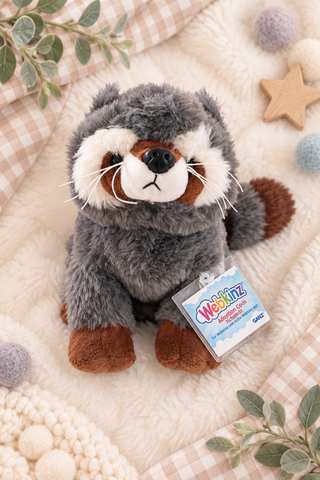 A plush raccoon toy from Webkinz sits on a soft cream blanket with decorative gingham fabric, pom-poms, leaves, and a wooden star. The raccoon is gray with white, brown, and black facial details and has a Webkinz hangtag attached.