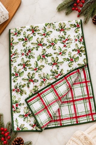 A folded holiday-themed kitchen towel or napkin with a white background, green holly leaf pattern, and red berries on one side, and a red and green plaid pattern on the reverse side with green piping. It is styled on a light marble surface with pine branches, red berries, and a wooden board.