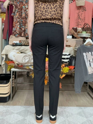 A woman viewed from behind wearing a leopard-print sleeveless top and black pants inside a clothing boutique. The store has racks of clothes, mannequins, and hanging garments in the background.