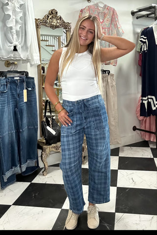 A woman modeling a pair of wide-leg blue plaid pants in a clothing boutique, paired with a white sleeveless top and beige sneakers.