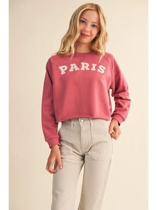A woman wearing a dusty rose pink cropped sweatshirt with 'PARIS' printed in white across the chest, paired with light beige jeans, against a solid light beige background.