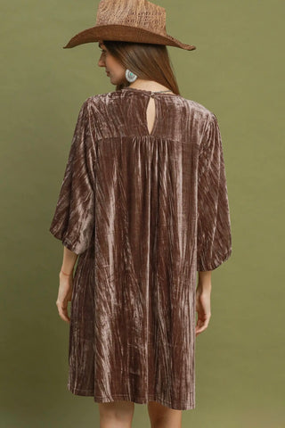 A woman viewed from the back wearing a brown crinkled velvet tunic dress with elbow-length sleeves and a small keyhole cutout at the back, paired with a brown hat, against an olive green background.