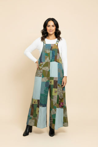 A woman modeling a pair of patchwork wide-leg jumpsuit overalls with green, blue, and beige tones, worn over a white long-sleeve top and black boots, against a solid beige background.