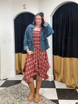 A woman stands in a fitting room wearing a red and white plaid strapless dress with a blue denim jacket and tan knee-high boots. The background features white walls, black and gold curtains, and a black-and-white checkered floor.