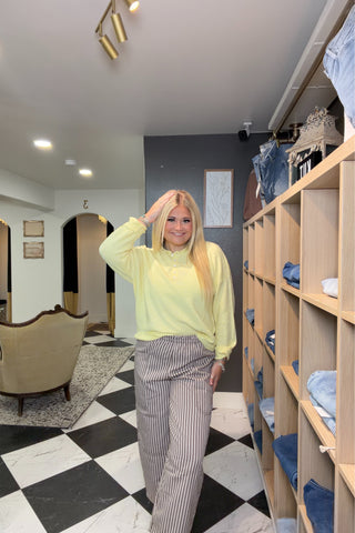 Woman in a bright yellow sweater and striped pants standing in a room with shelves and mirrors.