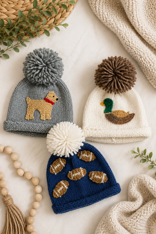 Three hand-knit baby beanies with pom-poms are arranged in a flat lay on a cream fabric background with decorative props. The left beanie is gray with a tan dog applique and a gray pom-pom.
