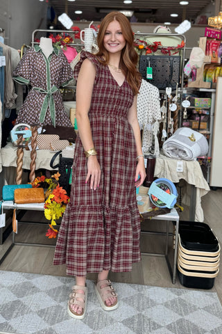A woman modeling a sleeveless, tiered plaid midi dress in a busy retail store setting. The dress features a deep V-neck and a rust-red and dark brown plaid pattern.