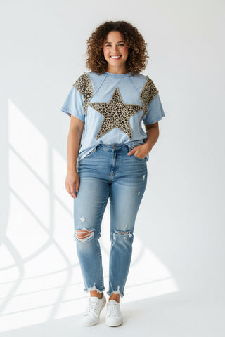 Light blue shirt with leopard print star and shoulder patches on a hanger against a brick wall.