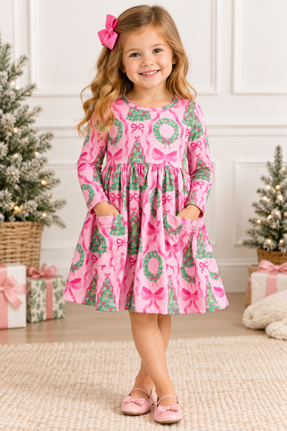 A young girl with a pink bow in her hair stands in a decorated holiday room wearing a pink long-sleeve dress with a green and pink Christmas tree and wreath pattern. She is wearing pink shoes and standing on a beige rug with Christmas trees and wrapped gifts in the background.