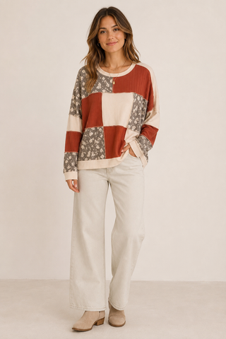 A woman modeling a long-sleeve patchwork knit sweater with rust red, cream, and gray floral blocks, paired with cream wide-leg pants and beige ankle boots, against a plain off-white studio background.