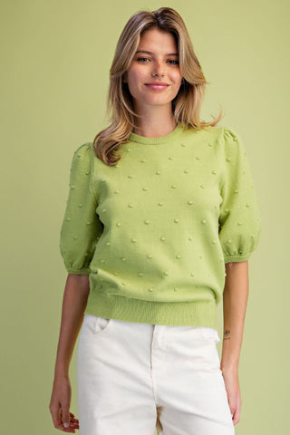 A woman wearing a light lime green short-sleeve knit sweater with a textured raised-dot pattern and white pants, standing against a solid light green studio background.