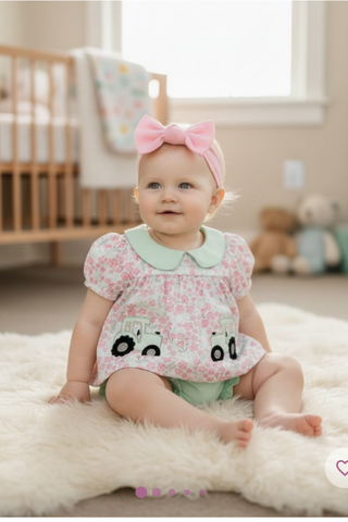 Baby sitting on a fluffy rug wearing a floral dress with black and white details and a pink headband.