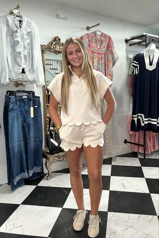 A woman modeling a matching cream short-sleeve top and shorts set with scalloped edges, standing in a clothing boutique with a black and white checkered floor.